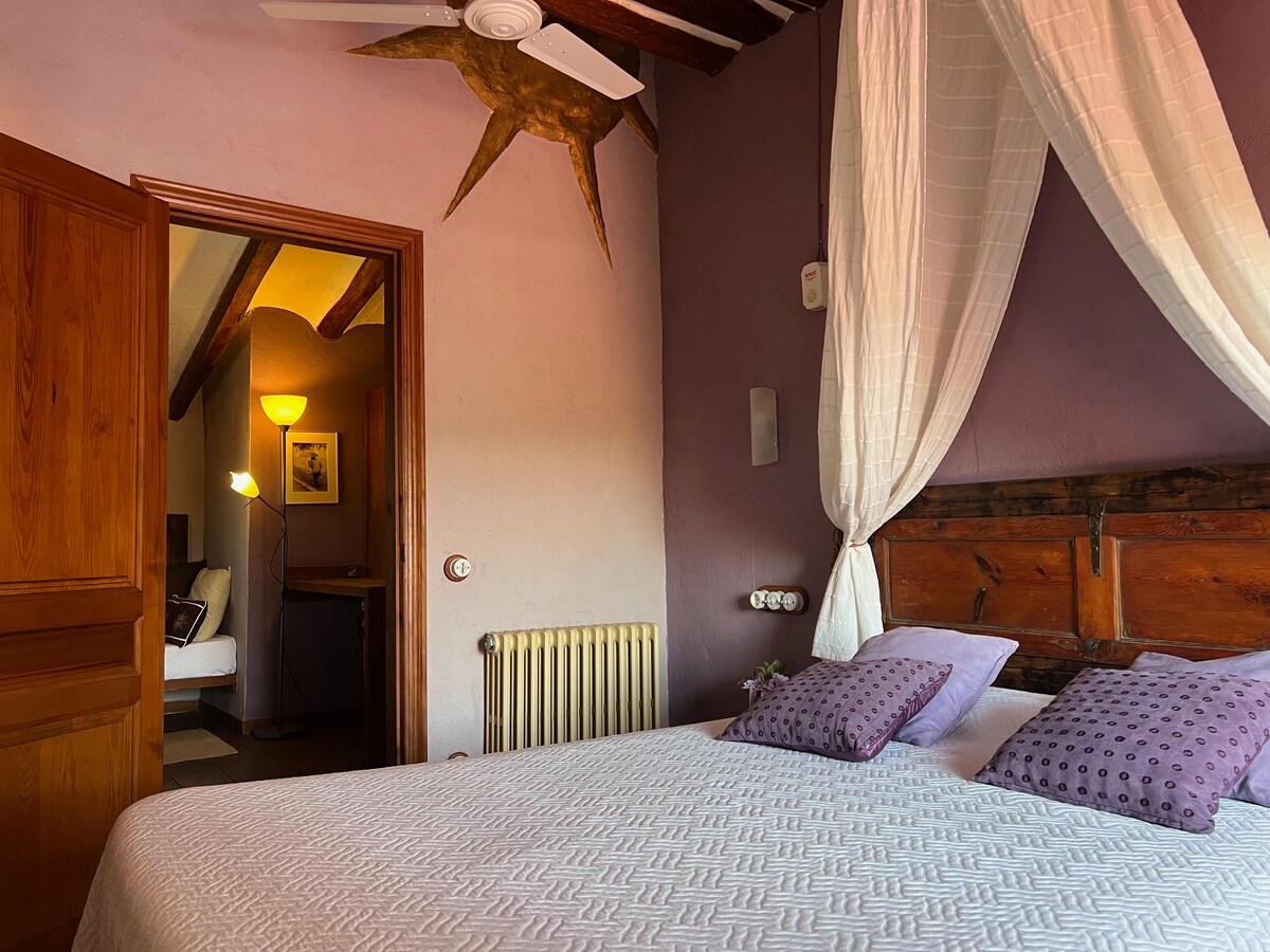 A restful bedroom is highlighted by a spacious bed with decorative pillows. Light fabric drapes gracefully from the ceiling, complementing the muted purple walls. A wooden door leads to an open space with additional furnishings, while a ceiling fan adds comfort to the inviting atmosphere.