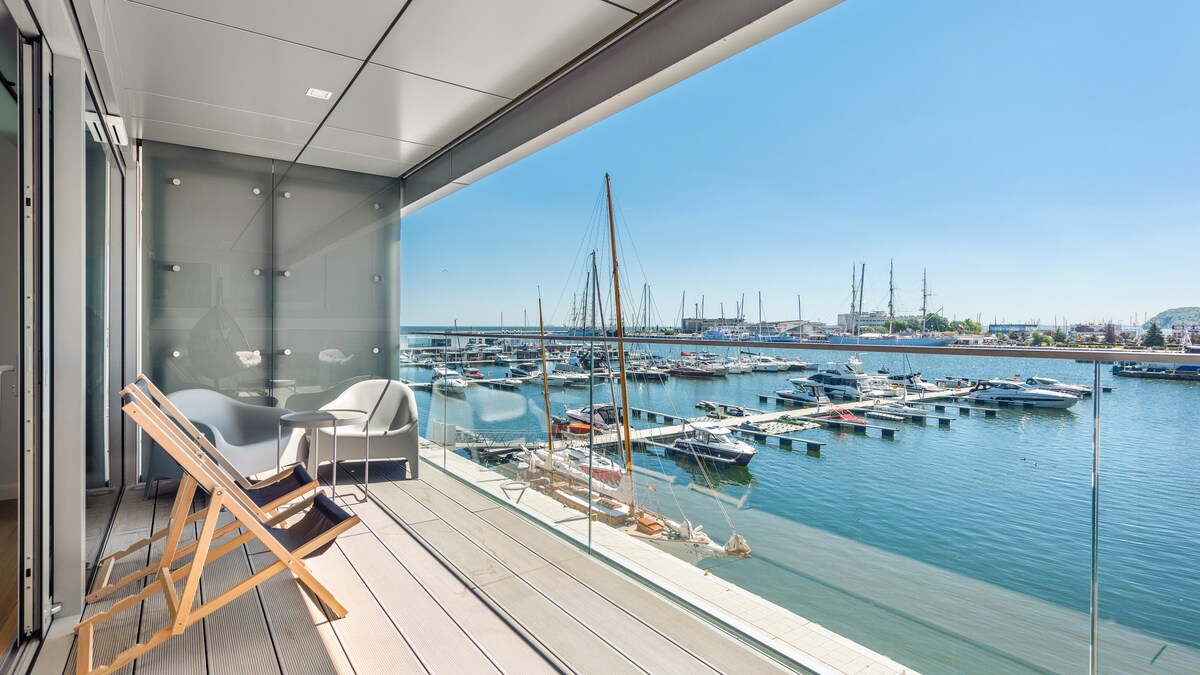 A private balcony features comfortable seating and a wooden lounge chair, overlooking a marina filled with boats. Direct views of calm waters and a clear blue sky create a serene outdoor space, with glass railings enhancing the open feel.