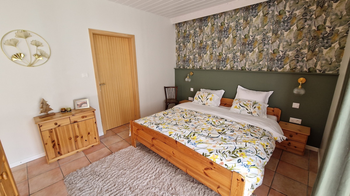 A bright bedroom features a wooden bed topped with floral-patterned bedding, complemented by matching bedside tables. A wall with nature-inspired decor adds a touch of color, while natural light filters through the space, highlighting a cozy chair and decorative elements on the dresser.