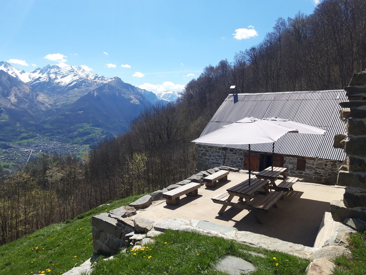 A stone chalet is set against a mountainous backdrop, featuring a wooden picnic area with benches. An umbrella provides shade over the seating area, surrounded by greenery and wildflowers. The scenic view encompasses the valley below and distant mountain peaks under a clear blue sky.