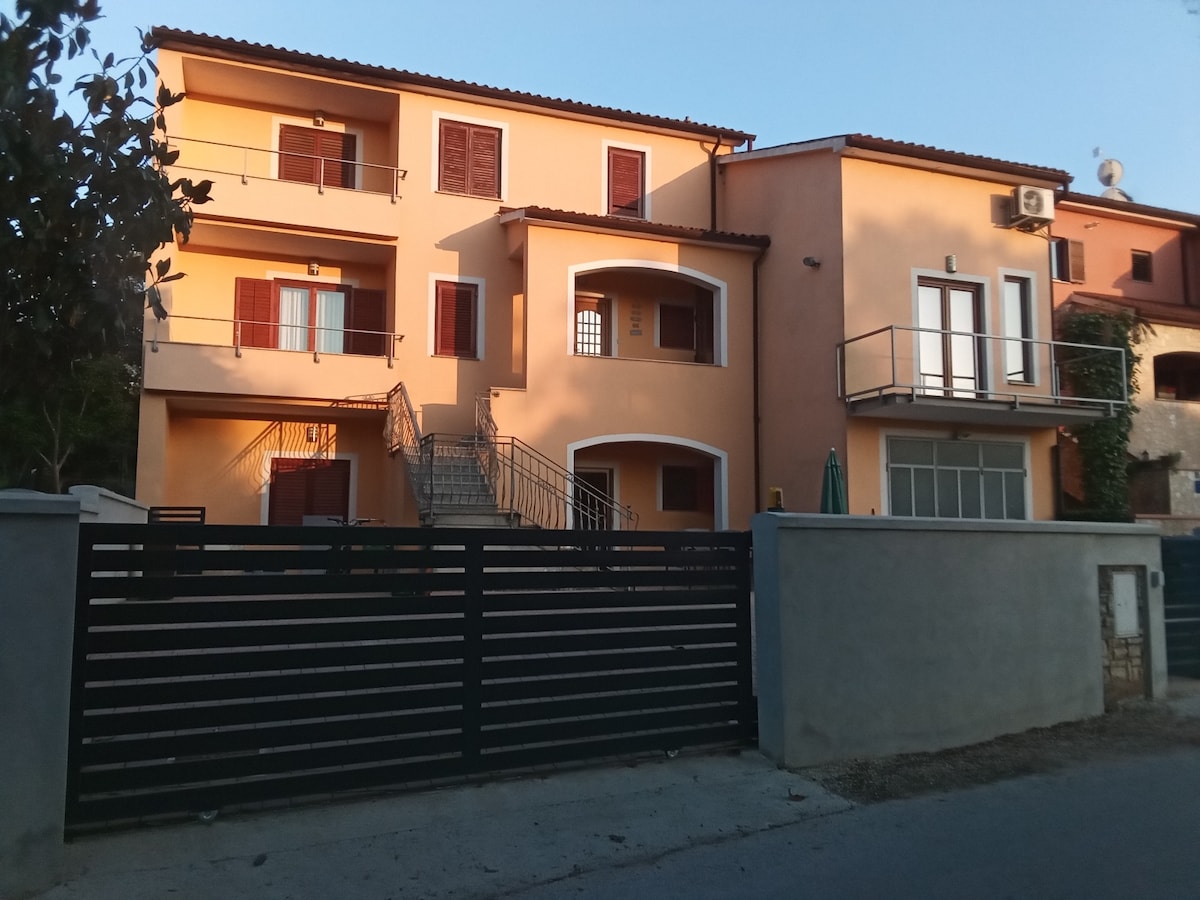 A three-story building is featured, constructed in a soft orange hue with contrasting brown shutters. A set of stairs leads to the upper levels, while a balcony above a large glass door offers outdoor space. A gated entrance provides access to the property.