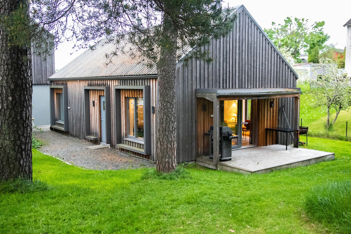 A modern wooden house is set against a backdrop of lush green grass and trees. Large windows provide views of the interior, while a covered porch offers seating options. The structure's contemporary design is highlighted by its sloped roof and natural wood finish.