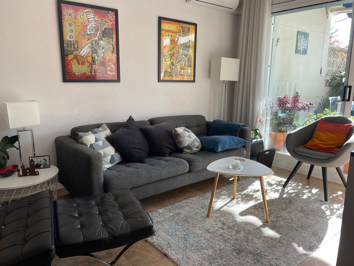 A modern living area is depicted, featuring a gray sofa adorned with various patterned pillows. A stylish coffee table sits in front, complemented by a light rug. Artwork hangs on the walls, and natural light floods in from a large window overlooking the terrace.