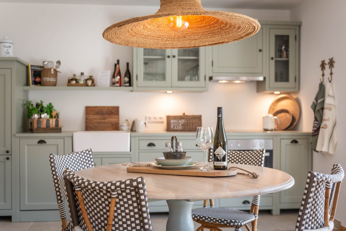 A light, airy kitchen is illustrated with muted green cabinetry and natural wood accents. A large round dining table, surrounded by checkered chairs, is centered beneath a spacious straw light fixture. Glassware and tasteful dishes are arranged on the table, enhancing a warm kitchen atmosphere.