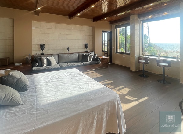 A spacious studio features a large bed with neutral bedding, accompanied by a comfortable sofa along one wall. Elegant wooden beams support the ceiling, while a large window offers views of the sea and natural surroundings. A cozy dining area with bar stools completes the layout.