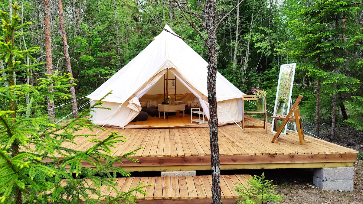 A spacious luxury glamping tent is positioned on a raised wooden deck surrounded by trees. The exterior features a light-colored canvas material, with a wooden railing leading to a small sitting area visible on the deck, offering a tranquil connection with nature.