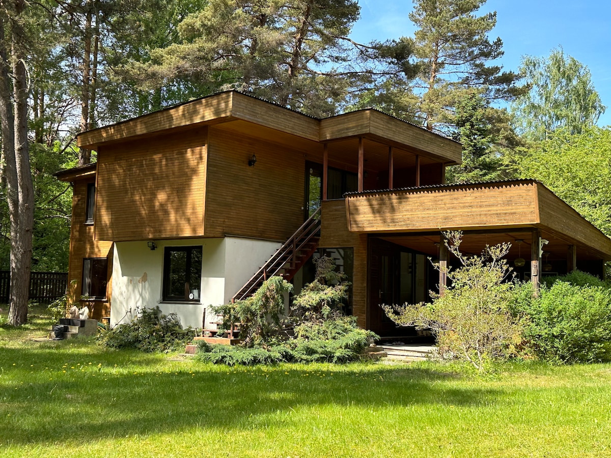 A two-story wooden house is surrounded by a lush garden, featuring a spacious wraparound deck. Large windows invite natural light into the interior. Trees provide shade and a sense of privacy, while the green lawn offers ample outdoor space for relaxation.