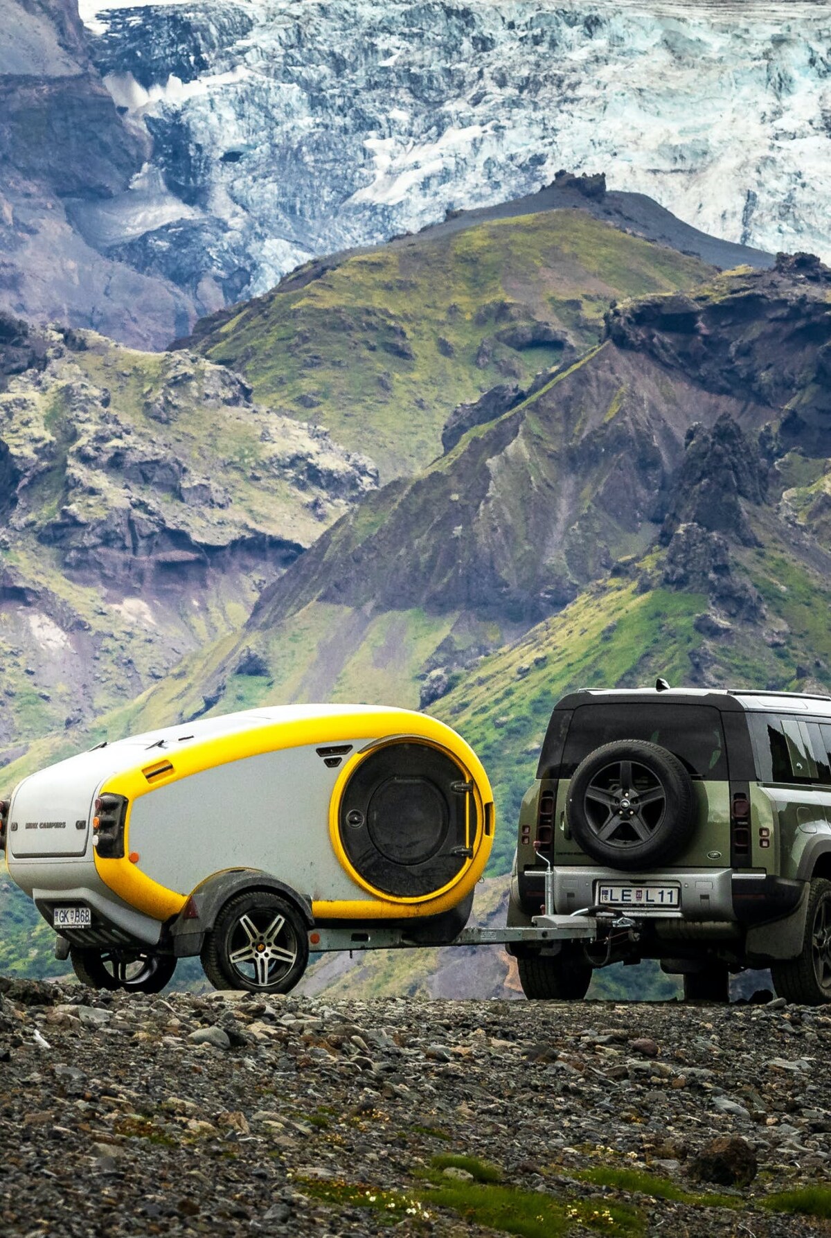 A compact trailer, featuring a sleek, rounded design with a yellow accent, is positioned next to an SUV on rocky terrain. A dramatic backdrop of lush green mountains and glacier-capped peaks creates a striking natural setting.