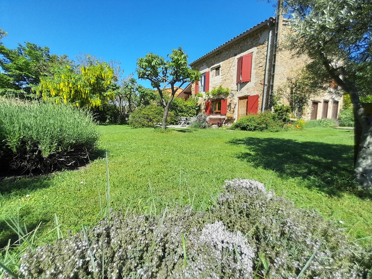 A charming stone house with red shutters is framed by lush greenery in the garden. Several trees and flowering plants add to the outdoor tranquility, while manicured grass creates an inviting space for relaxation and enjoyment of nature.