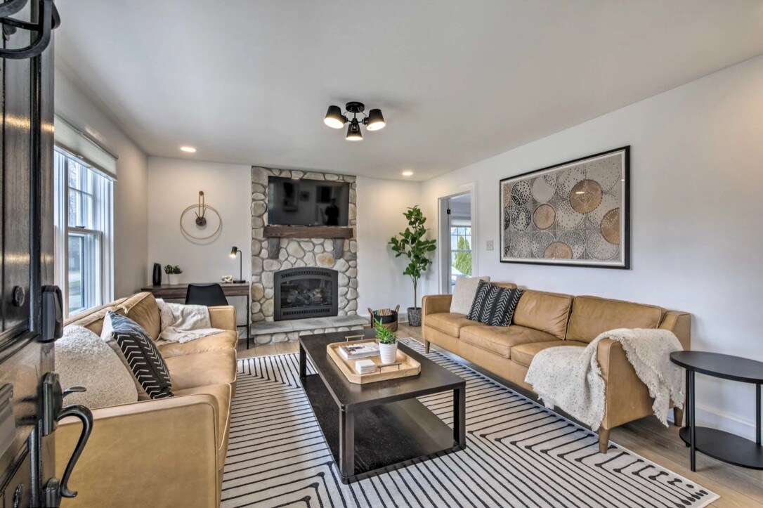 A welcoming living area is furnished with light brown sofas positioned around a dark coffee table. A stone fireplace serves as a focal point, while decorative wall art and a potted plant enhance the space. Natural light filters through the windows, illuminating the striped area rug.