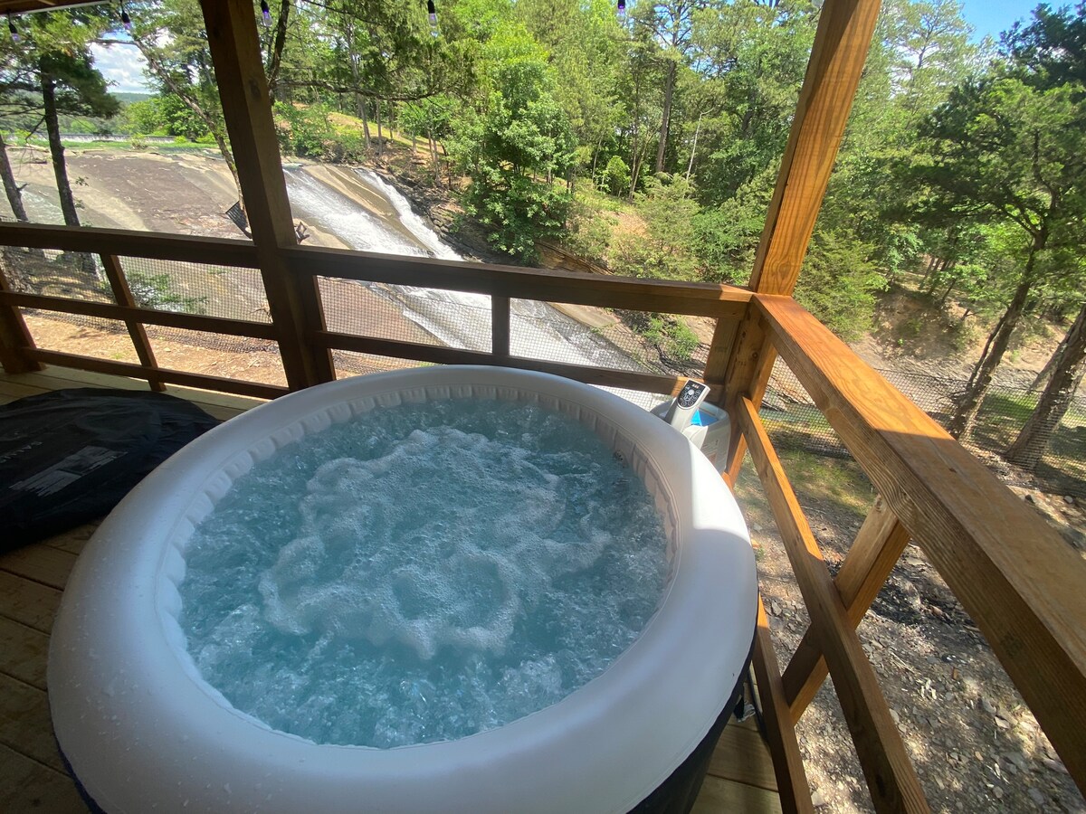 A hot tub with bubbling water is situated on a wooden porch. The backdrop features a picturesque view of a waterfall surrounded by lush greenery. Sunlight filters through the trees, creating a serene outdoor environment.