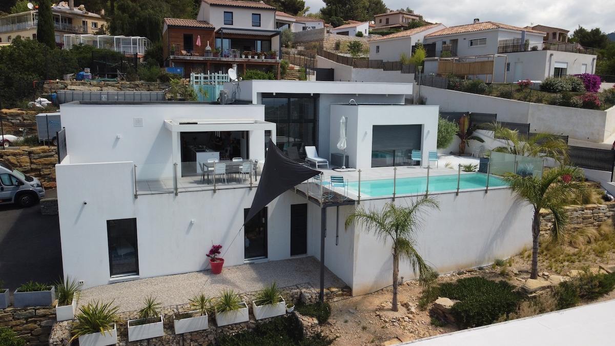 An architectural villa is showcased from an elevated angle, featuring a clean white exterior. A rectangular pool is visible, bordered by glass railings. Outdoor seating is arranged on the terrace, complemented by palm trees and a landscaped garden below.