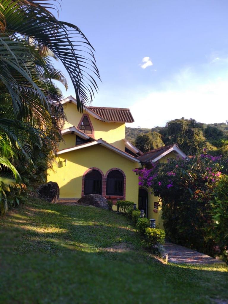 A charming two-story yellow house is set amid lush greenery, with vibrant purple flowers in the foreground. The building features arched windows and a sloped roof, surrounded by a well-maintained garden with natural stone pathways leading up the hillside.
