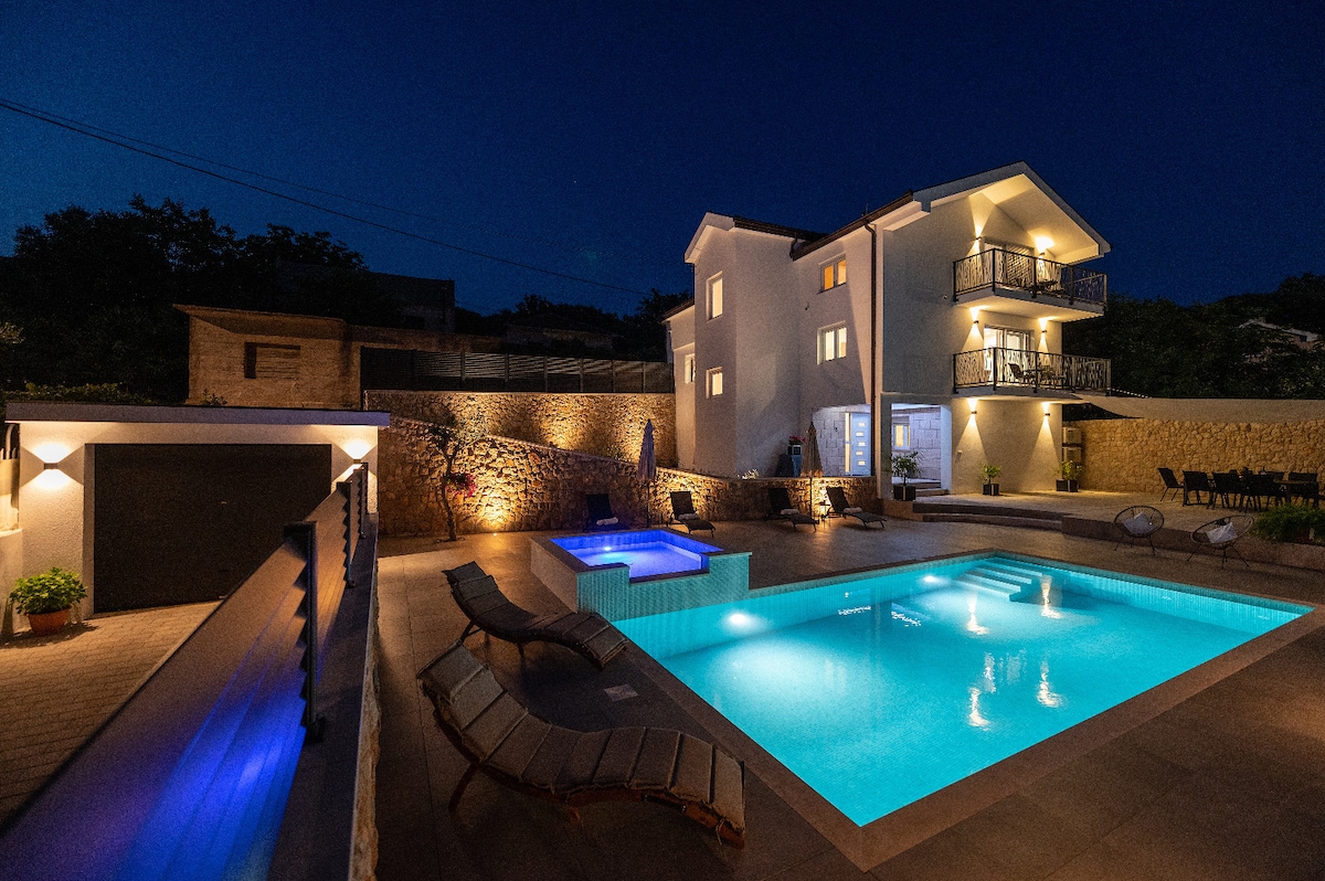 A newly constructed villa is illuminated at night, featuring a large outdoor swimming pool with clear blue water and surrounding stone deck. Lounge chairs line the poolside, while the three-story structure showcases multiple balconies. The garage is visible, and the ambiance is peaceful and serene.