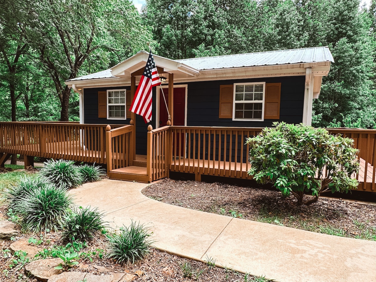The cabin's exterior is complemented by a welcoming wooden ramp and deck. An American flag is displayed prominently. Greenery surrounds the pathway leading to the entrance, with shrubs and trees providing a natural backdrop.