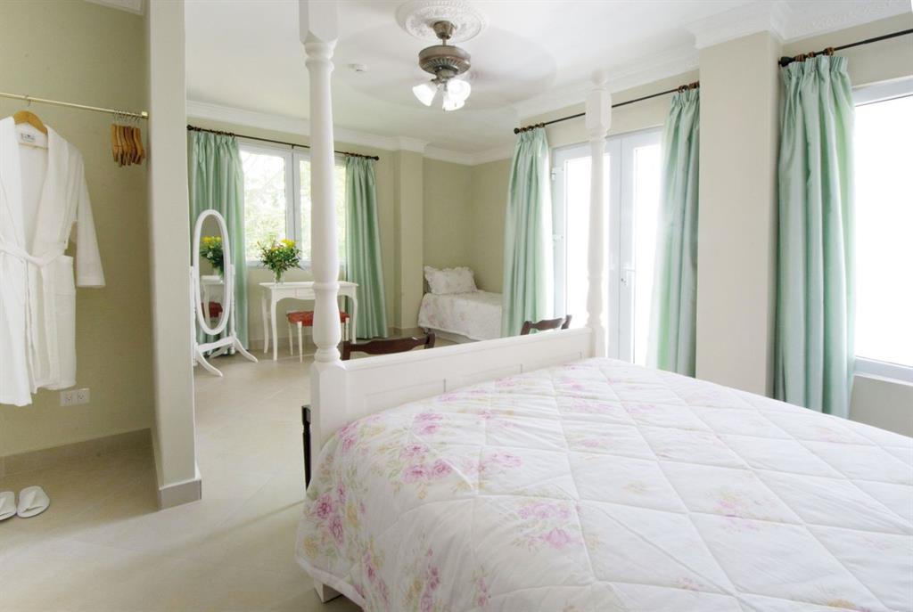 A light-filled room features a spacious queen-size bed with floral bedding. A seating area with a twin bed and a table is visible. Large windows draped with soft curtains allow natural light to enter. A robe hangs on a nearby rack, complementing the relaxing atmosphere.