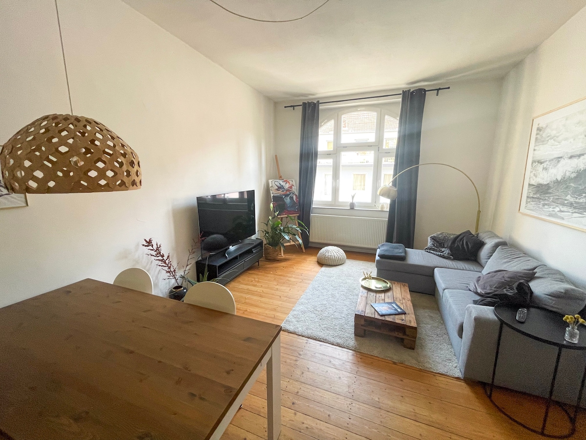 A bright living area is showcased, featuring a spacious gray sofa with decorative pillows and a coffee table made of natural wood. A TV is positioned on a low stand, and a large window allows natural light to illuminate the room. A dining table with white chairs is also visible.
