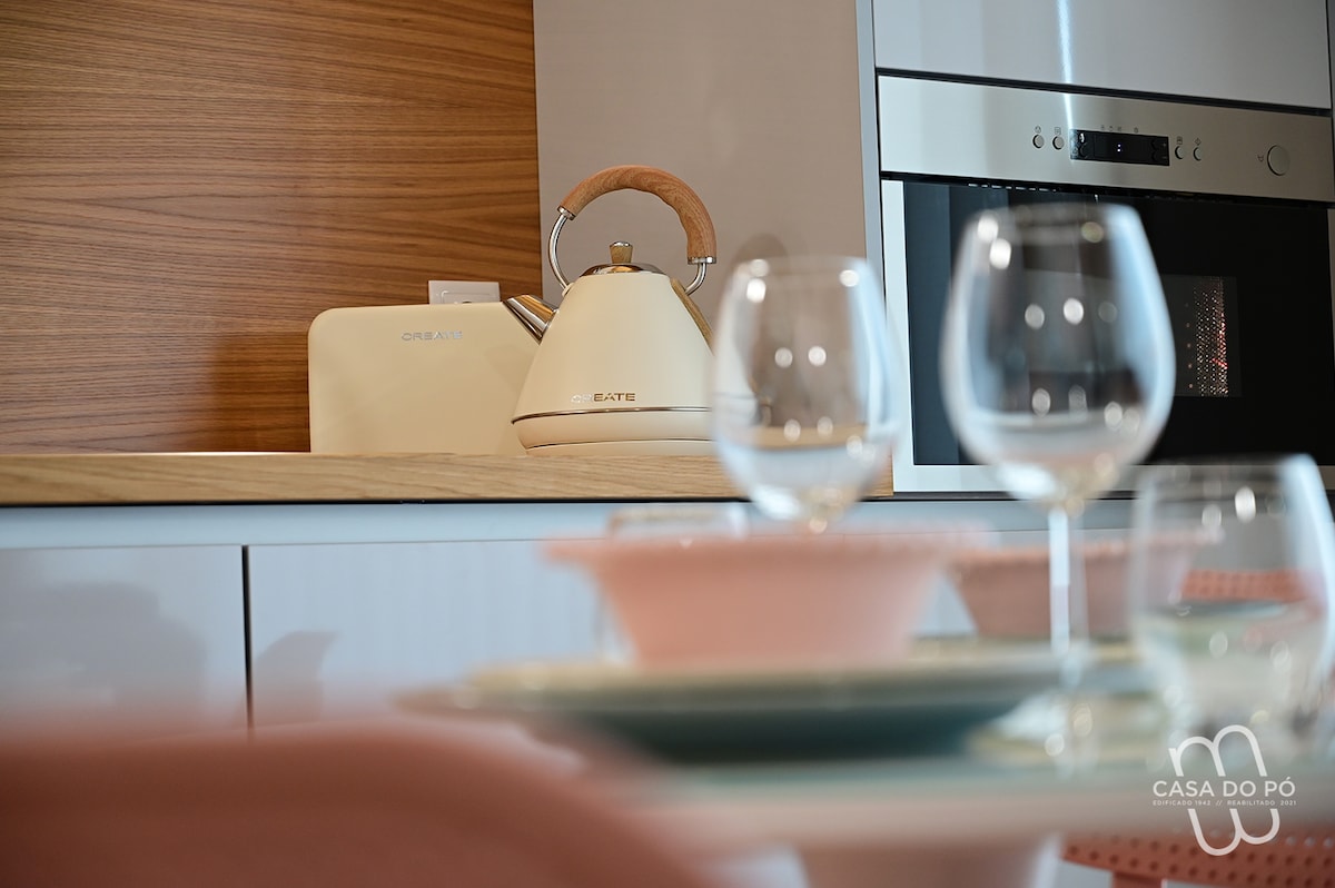 A modern kitchen is featured with wooden accents and sleek cabinetry. A light-colored kettle sits on the counter beside a retro-style toaster. A dining table is set with elegant glassware and pastel dishes, inviting guests to share meals in a stylish environment.