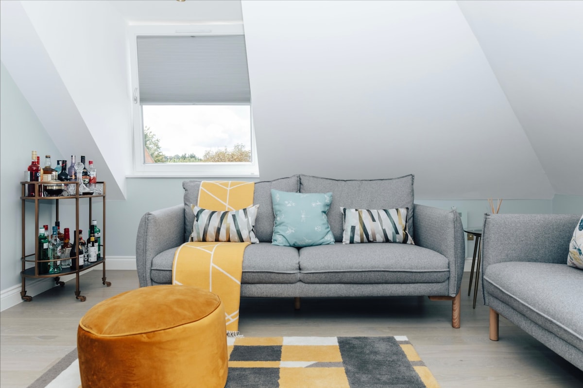 A well-appointed lounge area features a light grey sofa adorned with patterned cushions and a yellow throw. An orange pouf complements the space, which includes a stylish bar cart and a large window that allows natural light to fill the room.
