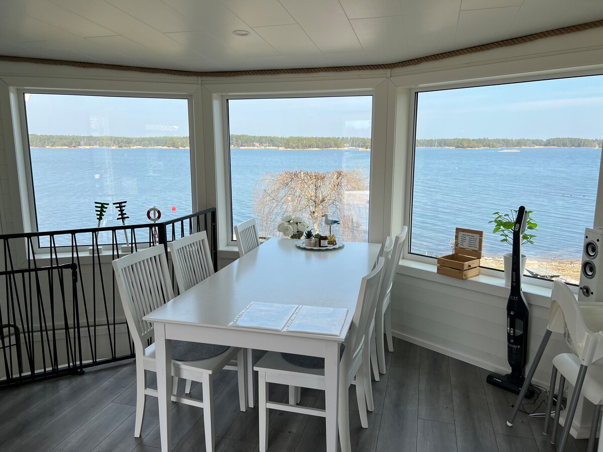A bright dining area features a large white table surrounded by six matching chairs, offering panoramic views through multiple large windows overlooking the serene lake. Soft natural light fills the space, enhancing the clean, modern aesthetic.