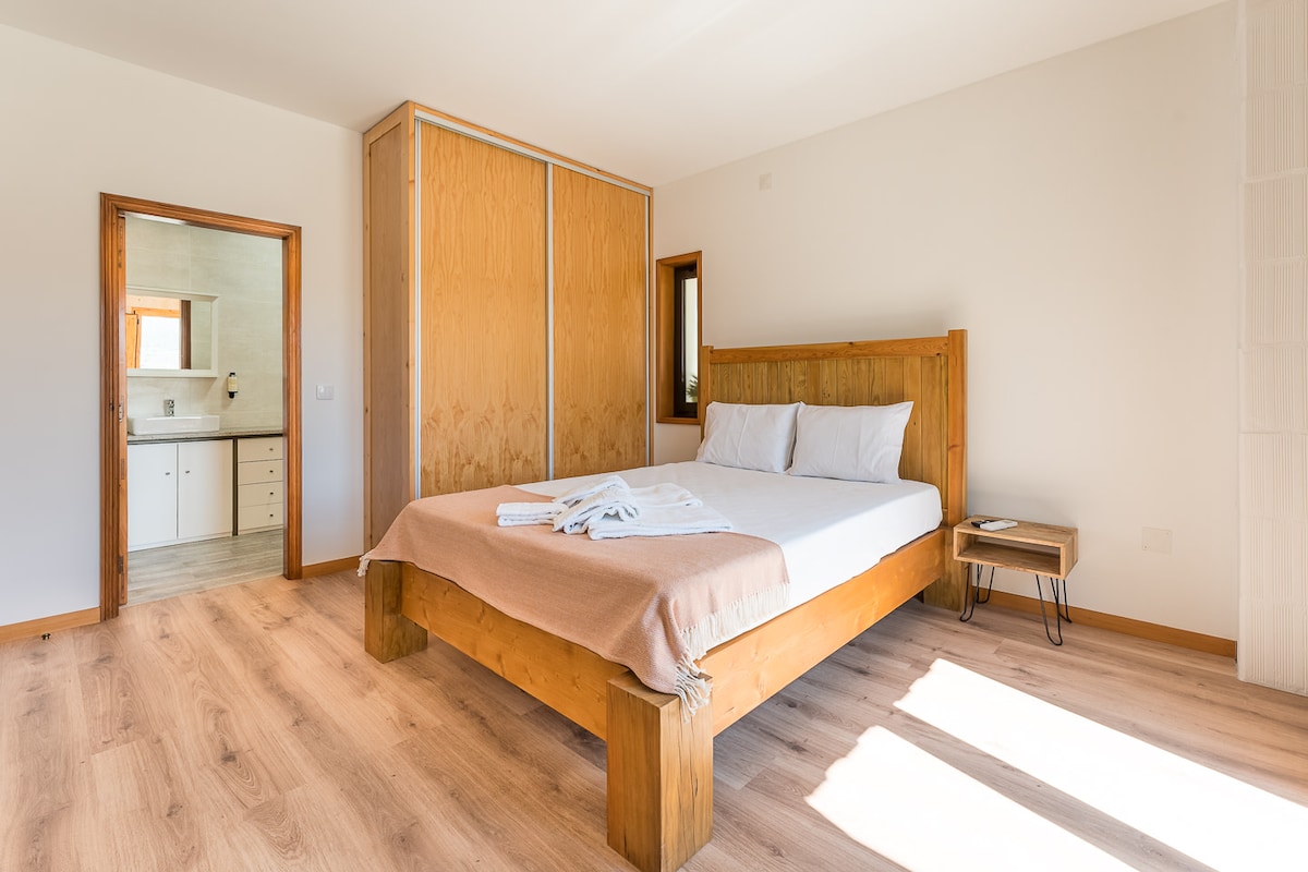 A spacious bedroom features a wooden bed topped with fresh linens and a soft blanket. Sunlight pours through nearby windows, illuminating the natural wood flooring. A bedside table and a built-in wardrobe complement the warm, neutral tones of the room.