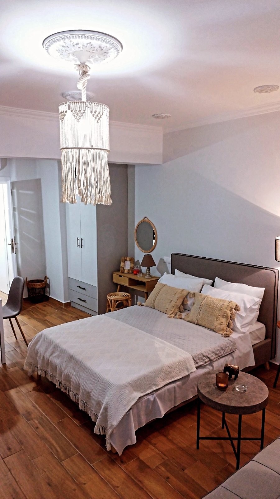 A modern studio features a spacious bed with neutral bedding and decorative pillows. A minimalist wooden table and chairs are present, along with a dresser and a circular mirror. Soft lighting is provided by a decorative chandelier, enhancing the overall design.