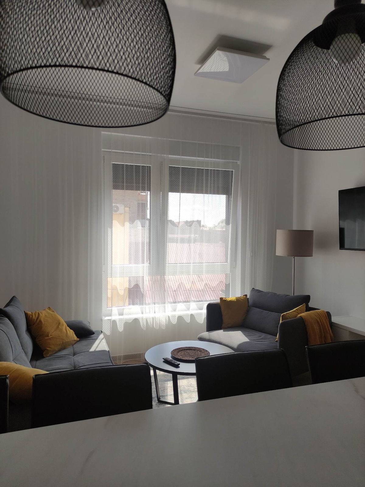 A cozy living area features a grey sofa adorned with yellow cushions, accompanied by a black coffee table. Large windows allow natural light to fill the space, complemented by sheer curtains. A lamp and a wall-mounted television add functionality to the environment.