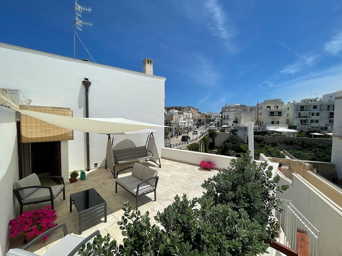 An outdoor terrace is depicted, featuring comfortable seating arrangements and a small table. Potted plants with vibrant pink flowers add color, while a sunshade provides a space for relaxation. A view of the surrounding area, including buildings and streets, is visible in the background.