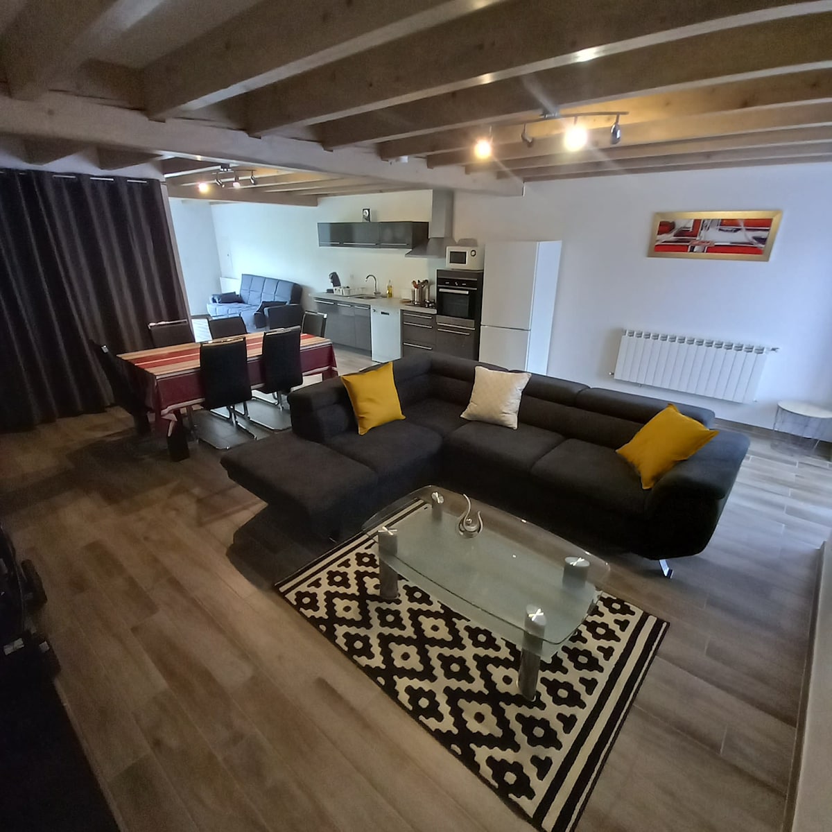A spacious living area features a dark sectional sofa with decorative cushions, paired with a modern glass coffee table. A dining table with black chairs is visible in the background, along with an equipped kitchen showcasing sleek appliances and a subtle color palette.