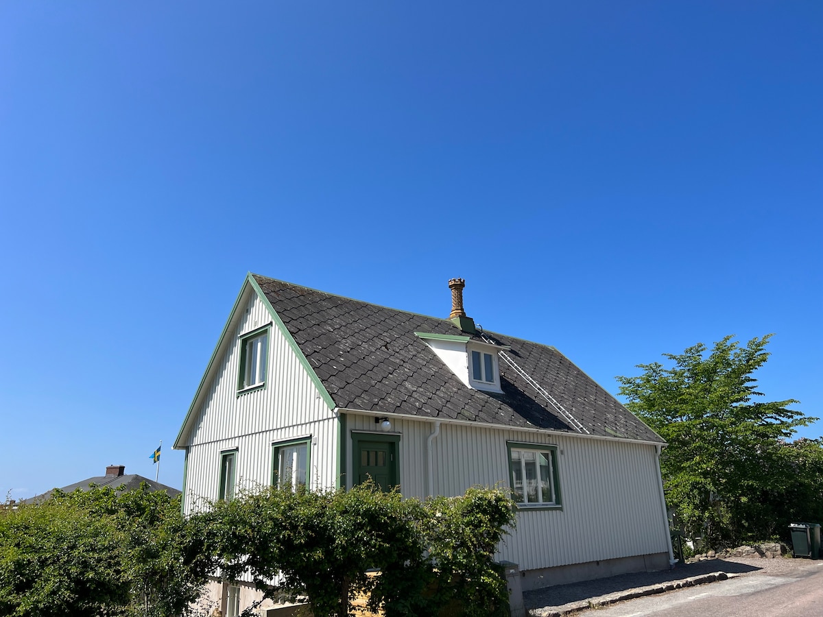 A charming, newly renovated house is presented with white siding and green accents. The sloped roof features a small chimney, and green foliage surrounds the entryway. A clear blue sky provides a bright backdrop, highlighting the inviting exterior.