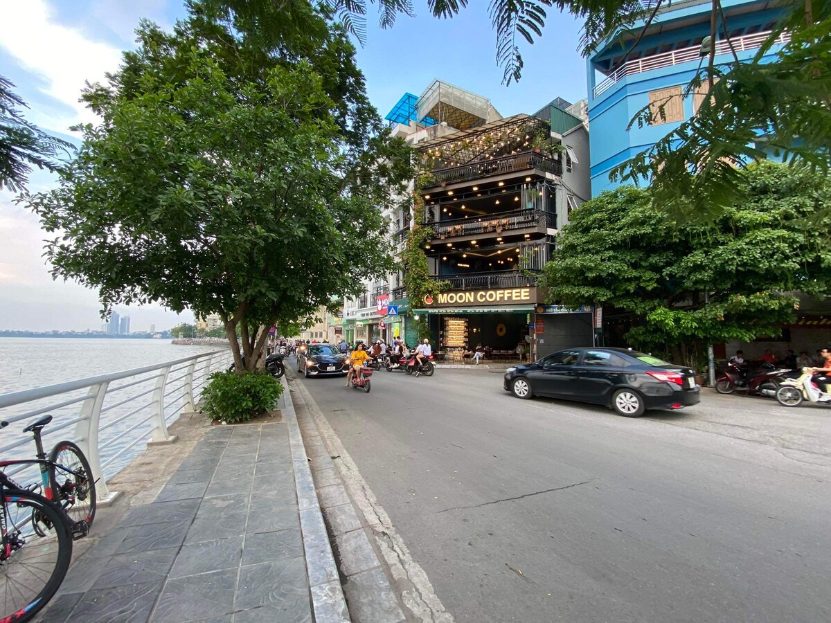 The image captures a bustling street scene along the West Lake, featuring a coffee shop with a prominent sign. Lush green trees line the sidewalk, and bicycles are parked nearby. Motorbikes and cars are visible on the road, contributing to the vibrant atmosphere.