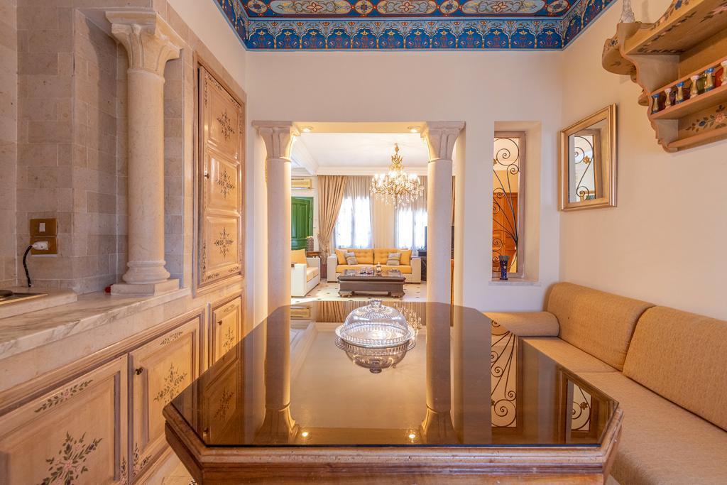 A dining area features a polished table with intricate details, surrounded by cushioned seating along the wall. Above, a beautifully patterned ceiling adds a touch of elegance. An archway leads into a spacious living area, softly illuminated by a chandelier.