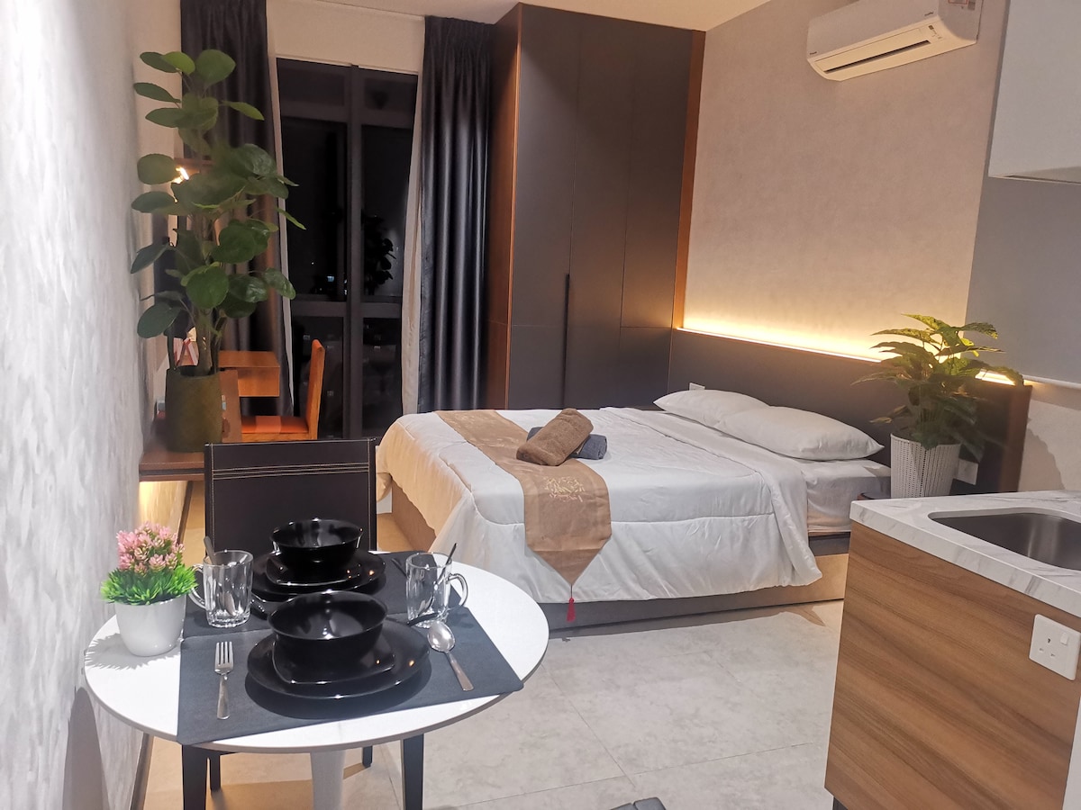 A modern studio room features a spacious bed with neutral bedding, complemented by sleek wooden furnishings. A dining table is set with tableware, while a kitchenette with a sink is included. Green plants add a touch of freshness, along with large windows allowing natural light.