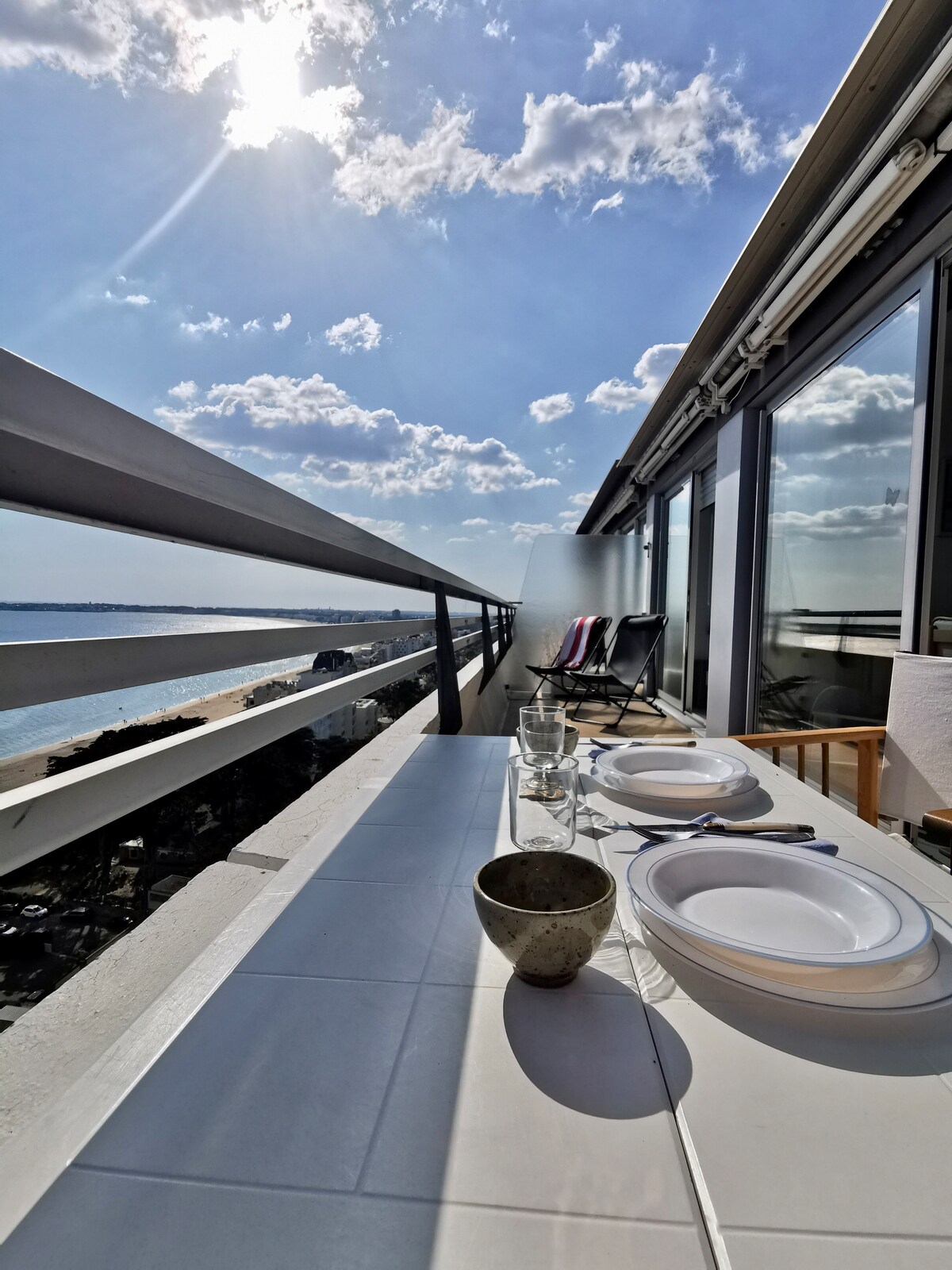 A spacious terrace features a dining table set with plates and cutlery, overlooking a stunning waterfront view. Two sun loungers are positioned alongside, and natural light pours in from the clear blue sky, highlighting the relaxed outdoor dining atmosphere.