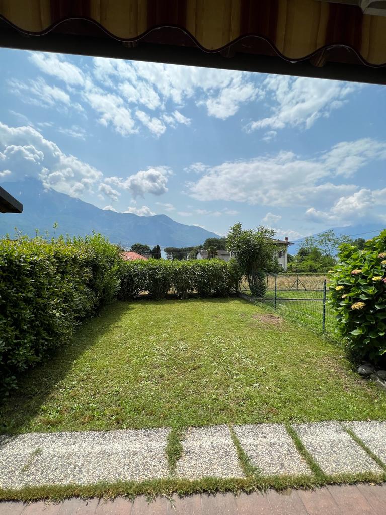 A private garden area is shown, featuring manicured grass bordered by hedges. Mountains are visible in the background under a bright sky with scattered clouds. A pathway leads from the house into the garden, providing a serene outdoor space.