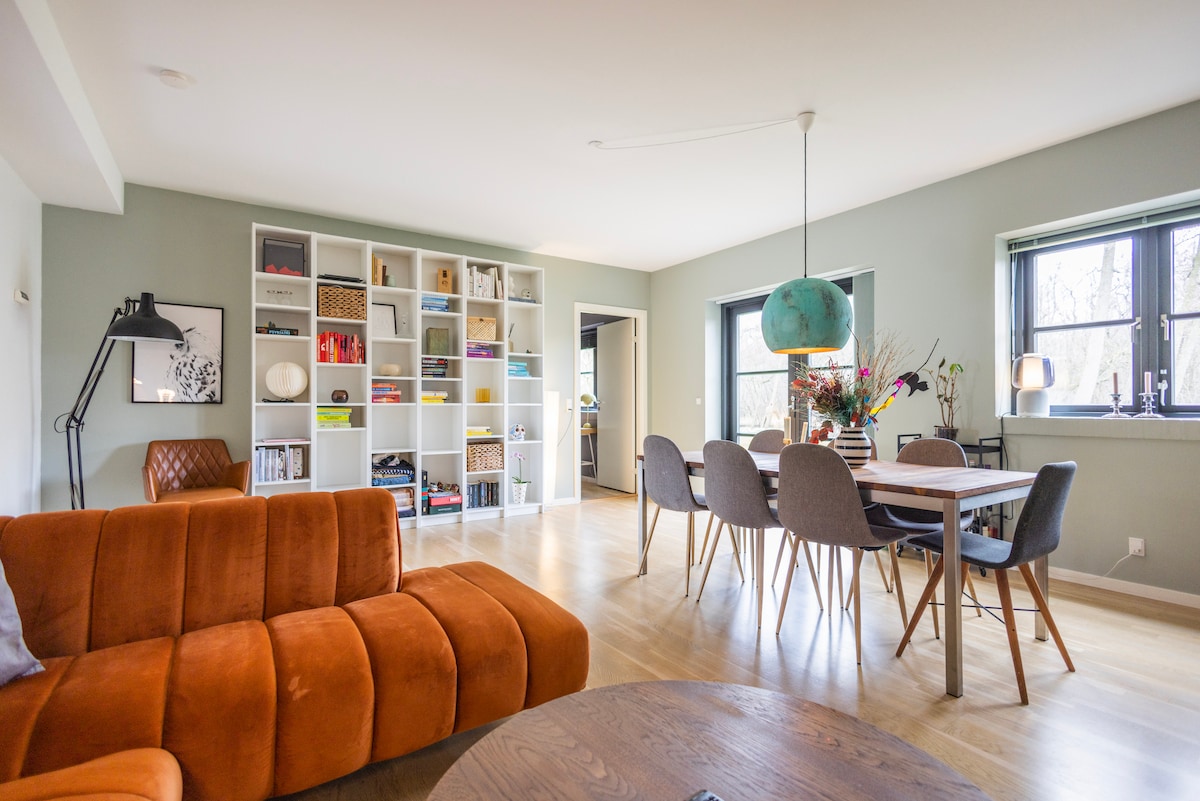 A modern living space features a warm orange sofa paired with gray dining chairs around a wooden table. A stylish bookshelf filled with various items is present against a light green wall, and large windows provide natural light to the inviting area.