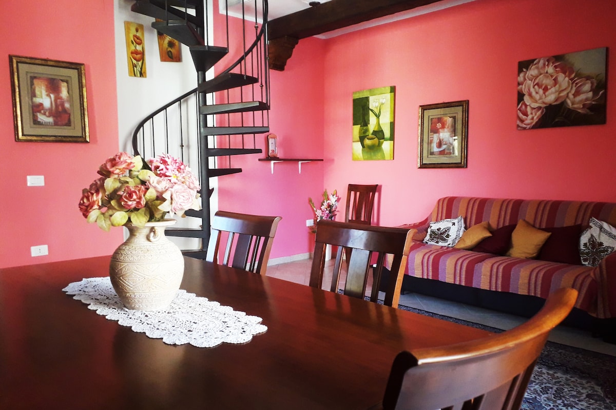 A dining area is furnished with a wooden table and several chairs, complemented by a decorative vase of flowers. Bright pink walls are adorned with floral artwork, while a spiral staircase leads to an upper level. A sofa with colorful cushions adds to the inviting ambiance.