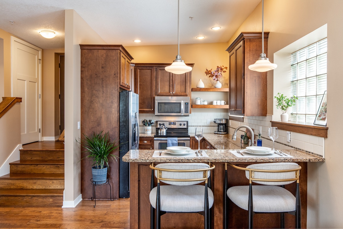 The kitchen features rich wood cabinetry and granite countertops, equipped with stainless steel appliances. Counter seating for two is highlighted by pendant lighting. A staircase is visible in the background, leading to the entrance of the apartment.