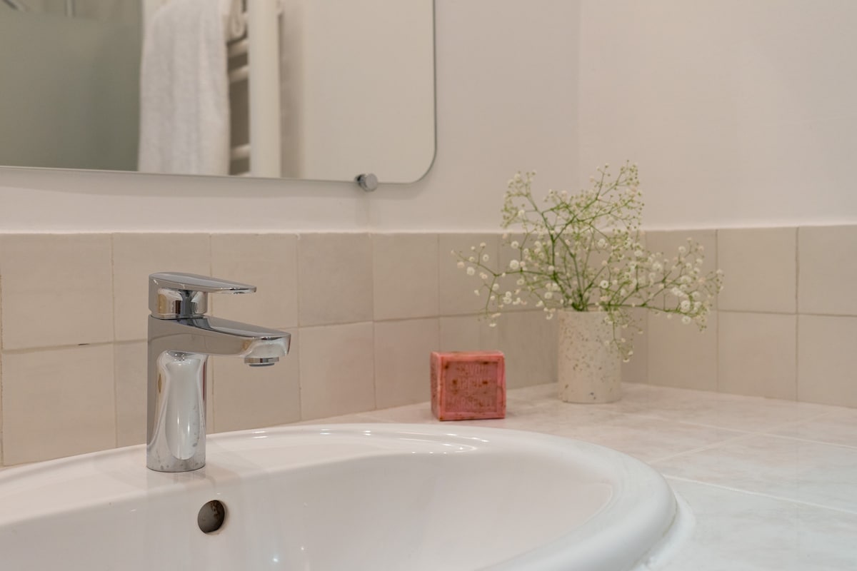 A bathroom sink area is shown, featuring a modern chrome faucet and a porcelain basin. A small vase with delicate white flowers and a decorative soap dish are placed nearby, contributing to a clean and tidy appearance. Soft natural light illuminates the space.