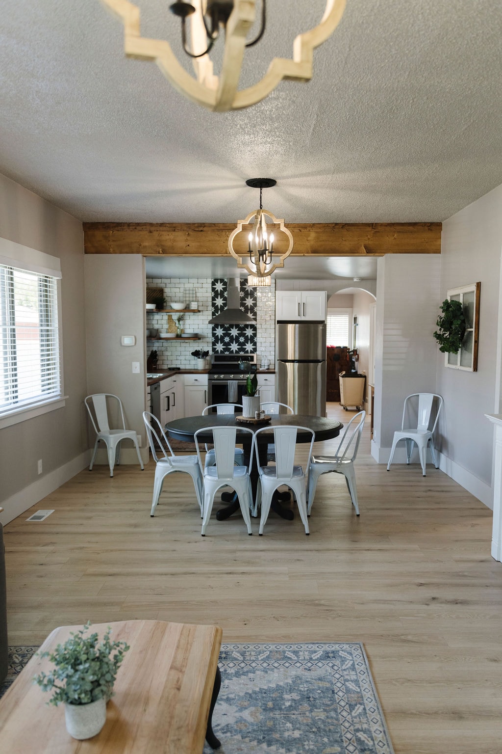 A dining area with a circular wooden table is surrounded by six metal chairs. Natural light enters through large windows, enhancing the light-colored walls and wood accents. The open layout connects to a modern kitchen featuring patterned tiles and stainless steel appliances.