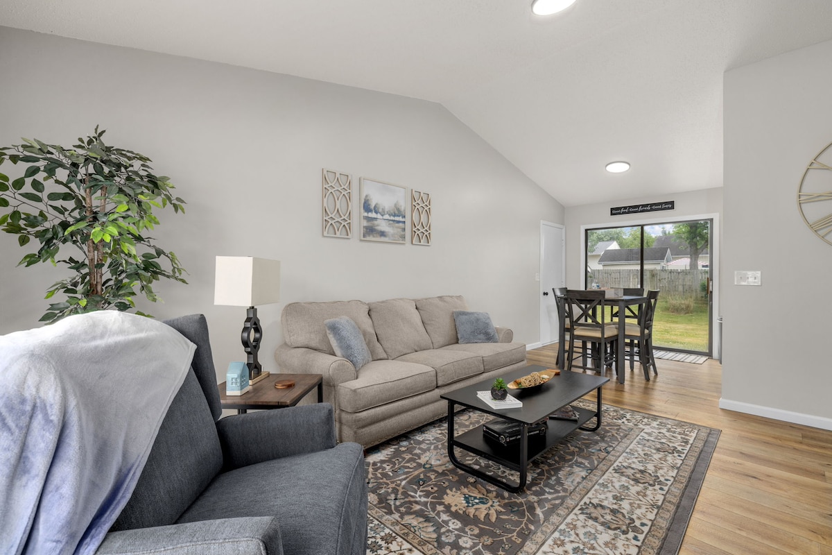 A bright living area features a comfortable sofa and armchair, arranged around a low coffee table. A large window offers natural light and views of the backyard. A dining area with tall chairs is positioned nearby, enhancing the open concept layout.