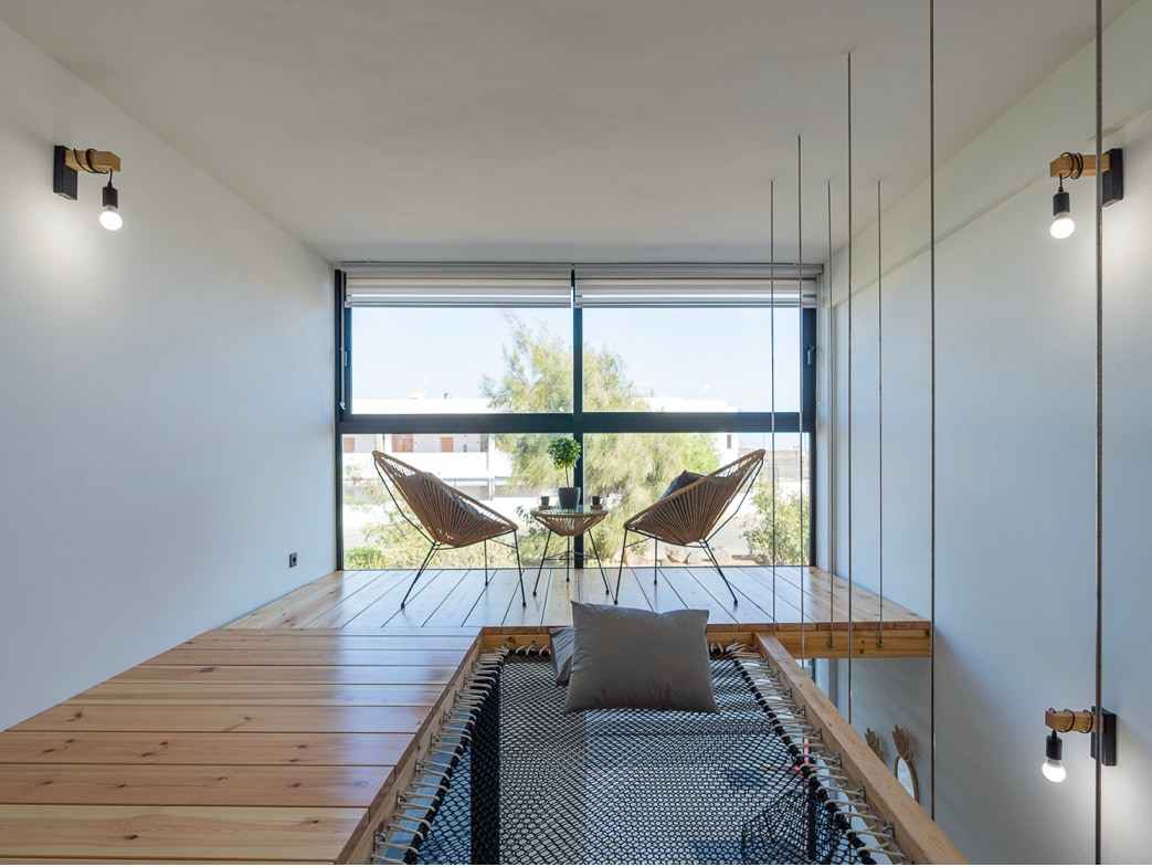 A bright loft area features a wooden platform with a large net and decorative pillow. Two stylish chairs face a window, inviting natural light and offering views of greenery outside. Simplistic lighting fixtures add to the modern aesthetic.
