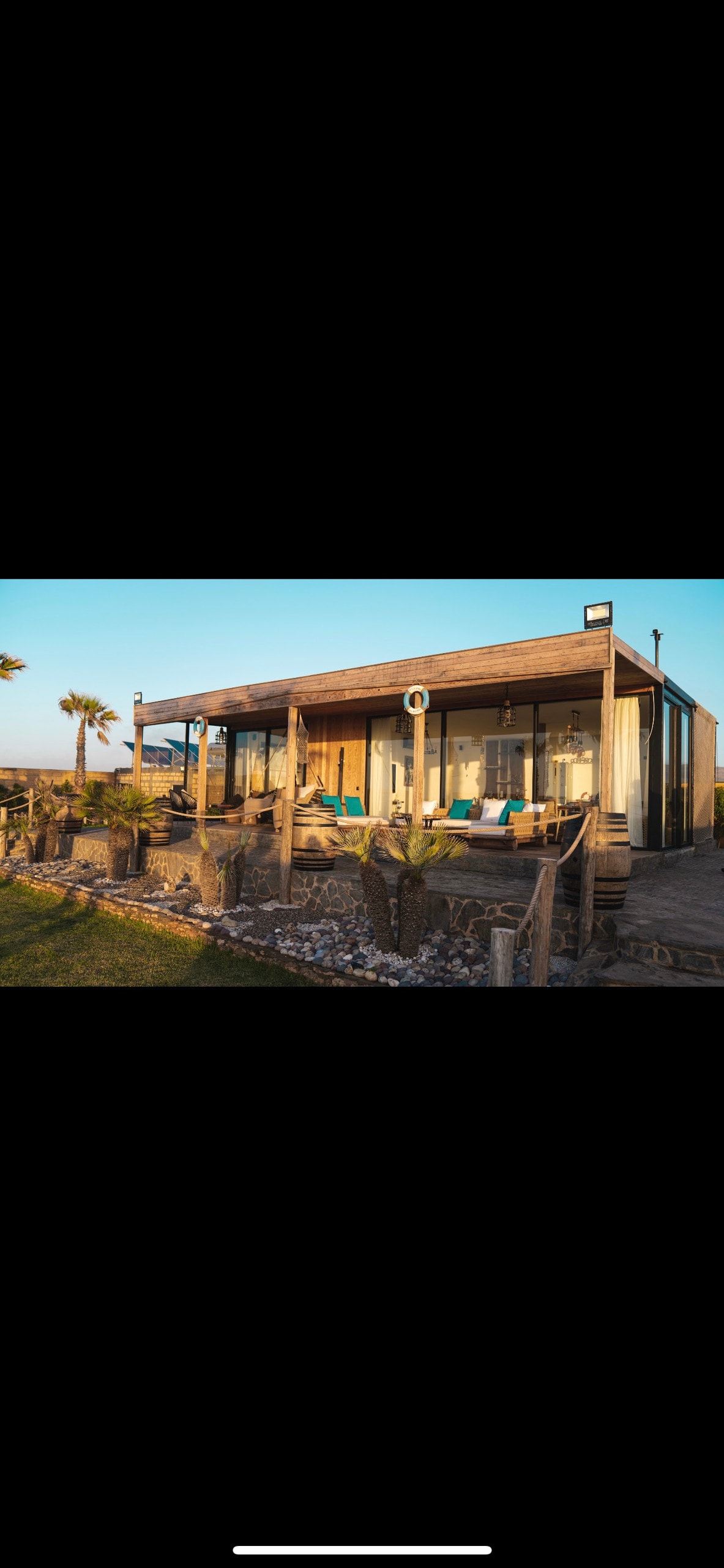 A charming coastal house is visible, featuring large deck areas with comfortable seating. Lush greenery and palm trees frame the exterior, while natural light enhances the wooden structure's inviting appearance. Decorative elements, such as seashells, are scattered in the foreground.