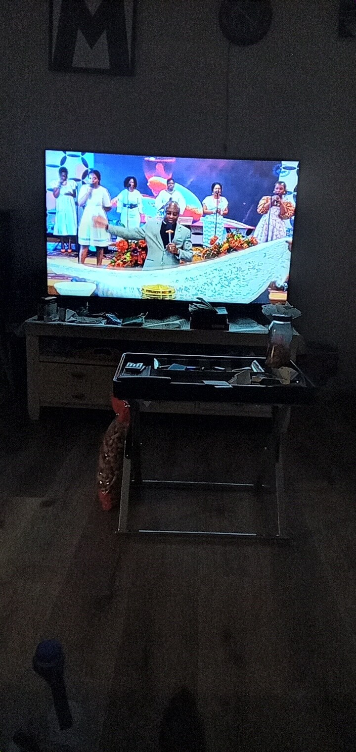A modern television is displayed on a console, showing a colorful performance featuring several individuals. A small table with various items is positioned in front of the television. The space is softly illuminated, highlighting the wooden floor.