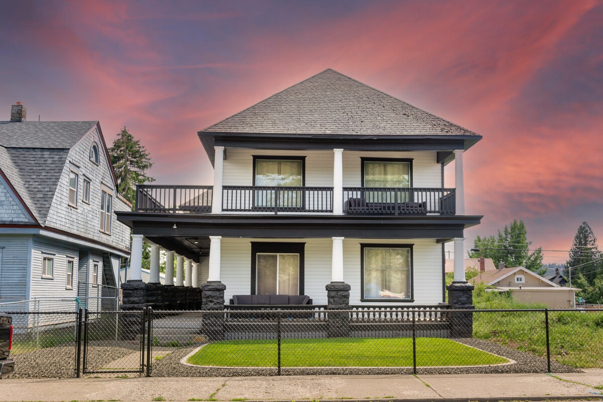 The exterior of a two-story house is depicted, featuring a covered balcony with railing on the second floor. Large windows allow natural light to enter. A manicured lawn with green grass is present in front, accompanied by a black fence along the property line.