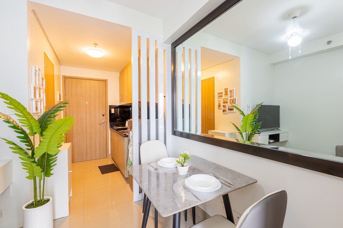 The dining area features a modern gray table with two chairs, complemented by a small vase of greenery. A mirror adds depth to the space, reflecting light. The kitchen is visible in the background, showcasing appliances and cabinetry, while the entrance is framed by wooden slats.