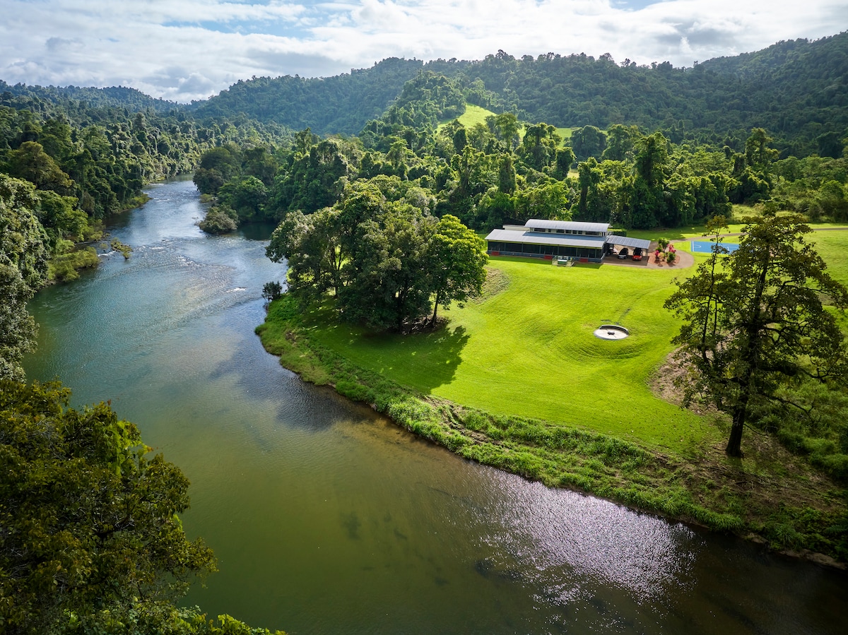 The image captures a scenic view of the Johnstone River, flanked by lush greenery and rolling hills. The Clubhouse at KC is visible alongside a cleared area with a hot tub and outdoor space, offering a tranquil setting surrounded by dense rainforest.