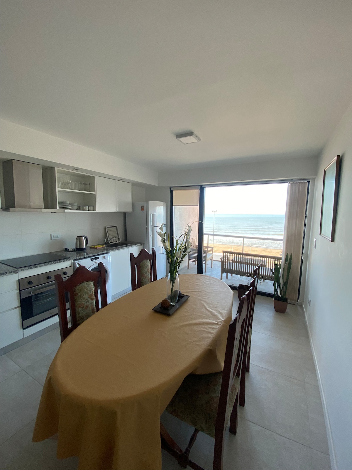 A modern dining area features a round table surrounded by six chairs, with a simple table setting. Large glass doors open to a balcony, offering views of the ocean. Natural light fills the space, highlighting the sleek kitchen cabinets and appliances.