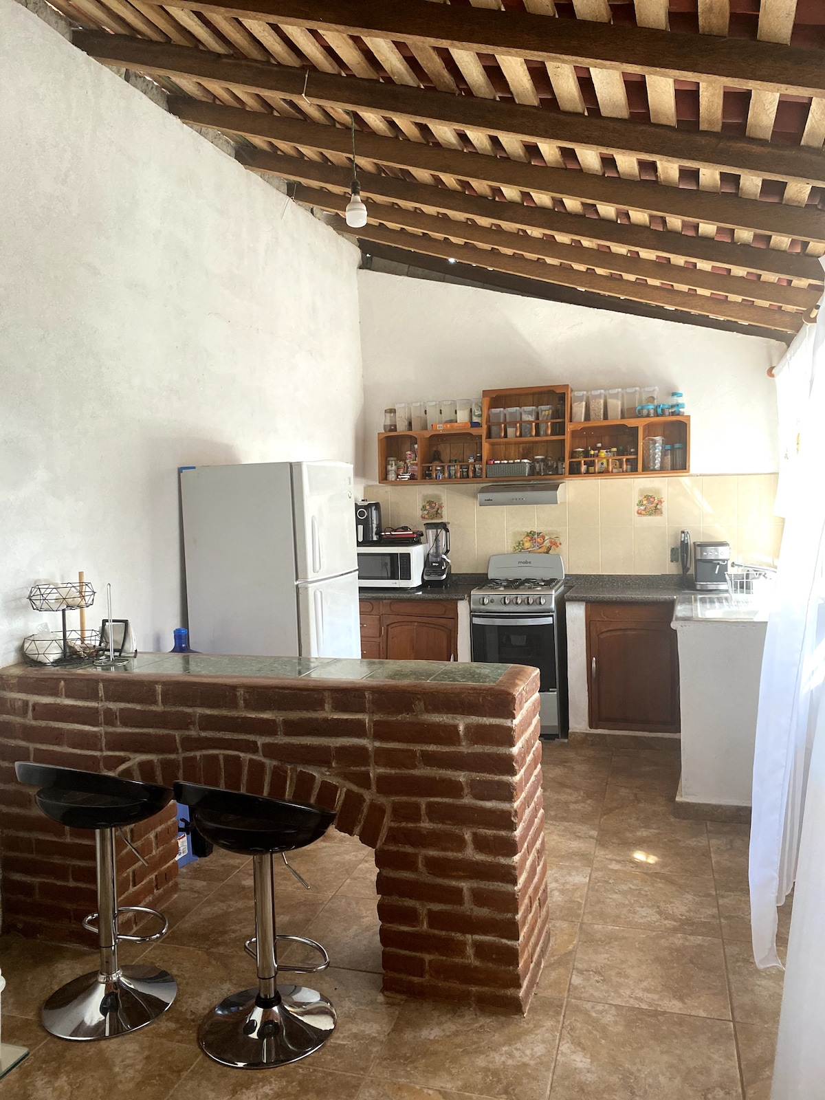 A compact kitchen features exposed wooden beams overhead and tiled flooring. A refrigerator and stove are present alongside a microwave, with ample countertop space. Two bar stools positioned at a brick counter provide seating, while various kitchenware is organized on open shelving.