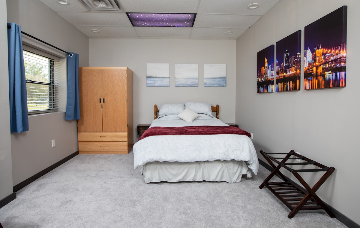 A spacious bedroom features a comfortable bed with a white comforter and a decorative pillow. A wooden wardrobe and a folding luggage rack are present. Three framed cityscape artworks adorn the walls, and natural light enters through a window with blue curtains.